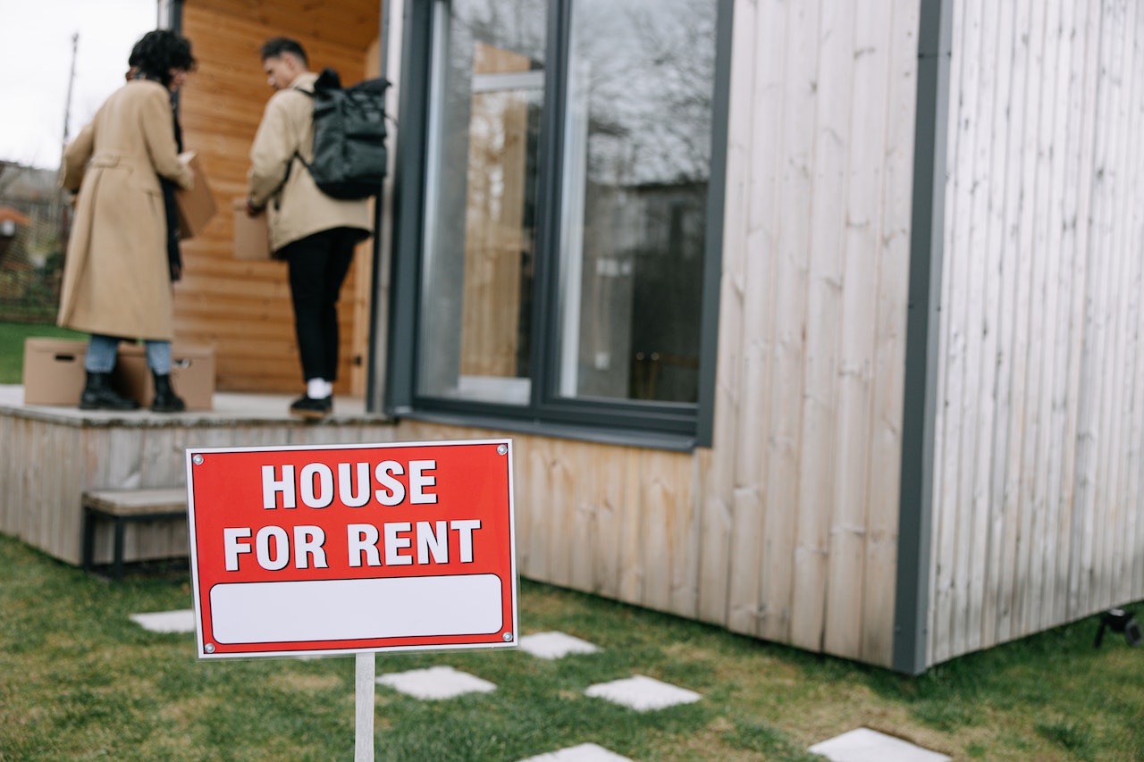 house for rent sign in front of a house. 2 people carrying boxes.